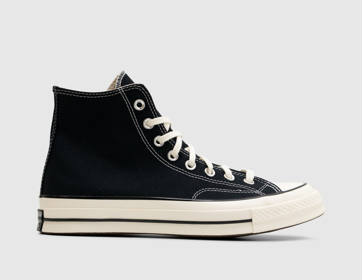 Converse Chuck 70 Hi. Side view of high-top canvas sneakers featuring a black canvas upper with contrast white stitching, silver eyelets, egret rubber toe cap, glossy egret rubber midsole with black pinstripe, and medial ankle patch positioning. 