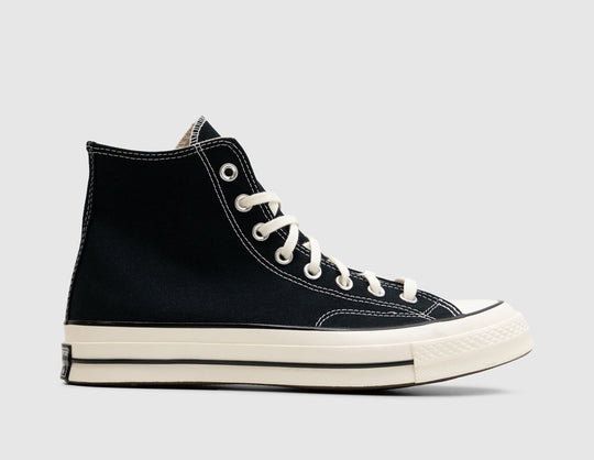 Converse Chuck 70 Hi. Side view of high-top canvas sneakers featuring a black canvas upper with contrast white stitching, silver eyelets, egret rubber toe cap, glossy egret rubber midsole with black pinstripe, and medial ankle patch positioning. 