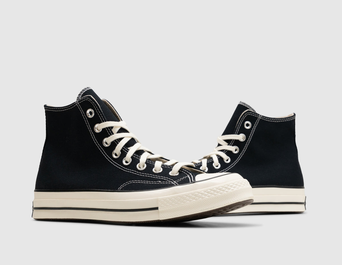 Converse Chuck 70 Hi. Three-quarter view of pair of high-top sneakers showing black canvas uppers, white stitching, silver metallic eyelets, egret laces, and vintage egret rubber outsoles. 