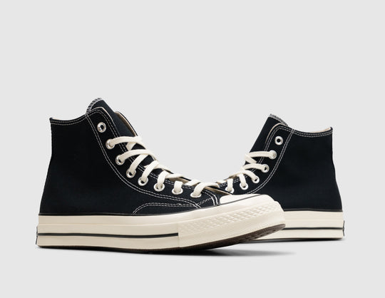 Converse Chuck 70 Hi. Three-quarter view of pair of high-top sneakers showing black canvas uppers, white stitching, silver metallic eyelets, egret laces, and vintage egret rubber outsoles. 