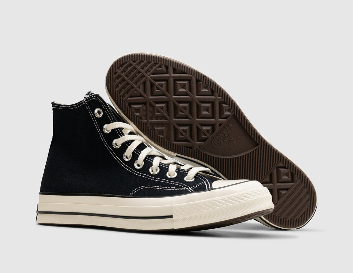Converse Chuck 70 Hi. Dynamic view of pair of high-top sneakers with the background shoe resting on its side to display the brown diamond-patterned rubber outsole behind the lateral profile of the front shoe. 