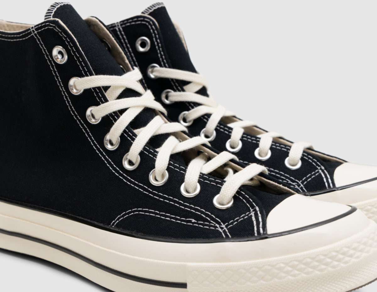 Converse Chuck 70 Hi. Close-up three-quarter view of the upper showing black canvas construction, white contrast stitching along the eyestay and toe box, silver eyelets, egret laces, and glossy egret rubber toe cap. 