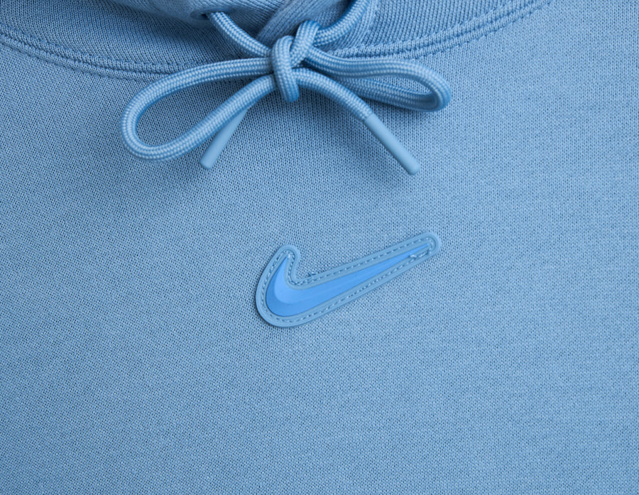 Nike NOCTA Fleece CS Hoodie. Close-up of work blue crossover hood with matching drawstrings and center gel Nike Swoosh logo. 