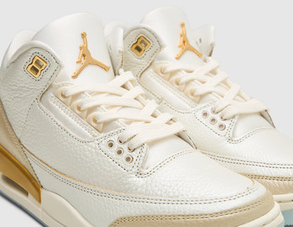 Jordan Women's 3 Retro. Close-up three-quarter view of sail tumbled leather upper showing metallic team gold Jumpman logo on tongue, gold top eyelets, and pale ivory laces. 