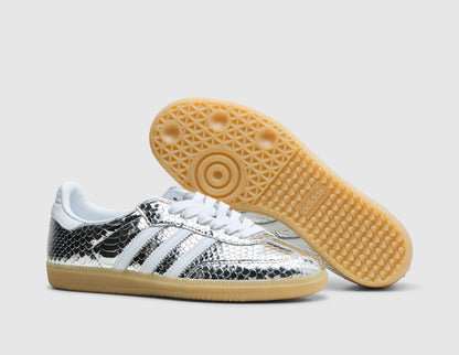 adidas Women's Samba. Three-quarter view of silver metallic snakeskin sneakers with one shoe propped to display the gum rubber tread outsole pattern. 