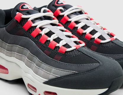 Nike Air Max 95 OG Big Bubble. Close-up three-quarter view of anthracite toe cap and mesh upper showing solar red ribbon lace loops, white laces, and solar red Air Max tongue oval logo. 