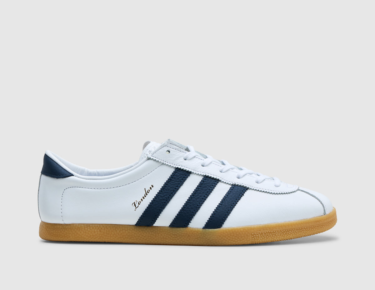 adidas London - size? Exclusive. Side view of footwear white leather sneakers featuring crew navy leather Three Stripes, crew navy heel tab, gold foil cursive "London" lateral branding, and low-profile gum 3 rubber sole unit. 