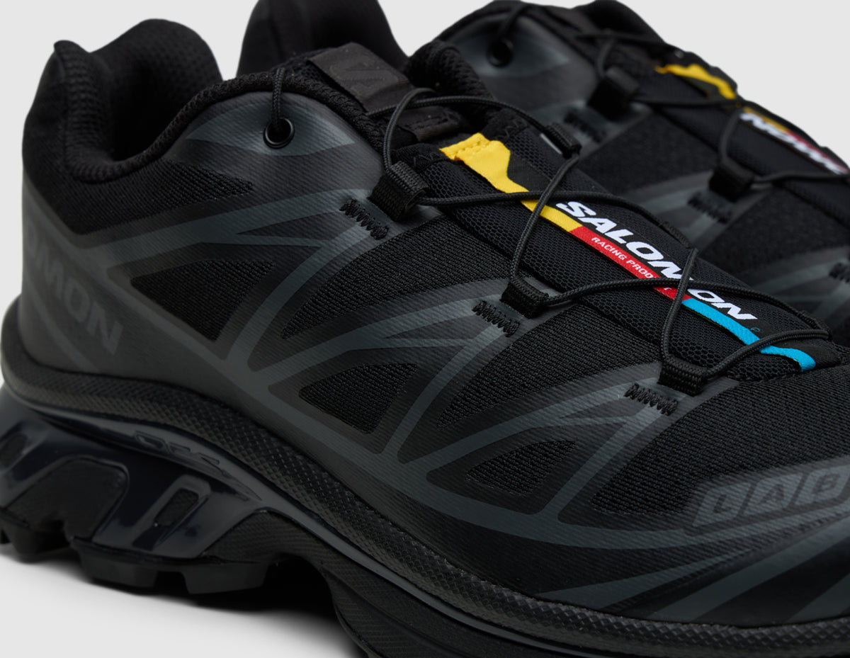 Salomon XT-6. Close-up three-quarter view of triple black trail running shoes showing Quicklace toggle system, colorful tongue tag, and toe cap branding. 
