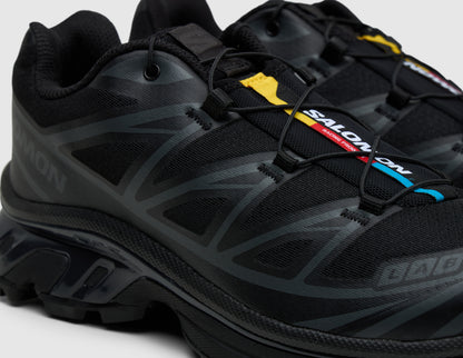 Salomon XT-6. Close-up three-quarter view of triple black trail running shoes showing Quicklace toggle system, colorful tongue tag, and toe cap branding. 